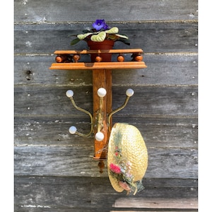 May include: A wooden coat rack with a shelf and four hooks. The rack has a small potted plant with purple flowers on the shelf. A straw hat with floral embellishments hangs on one of the hooks.