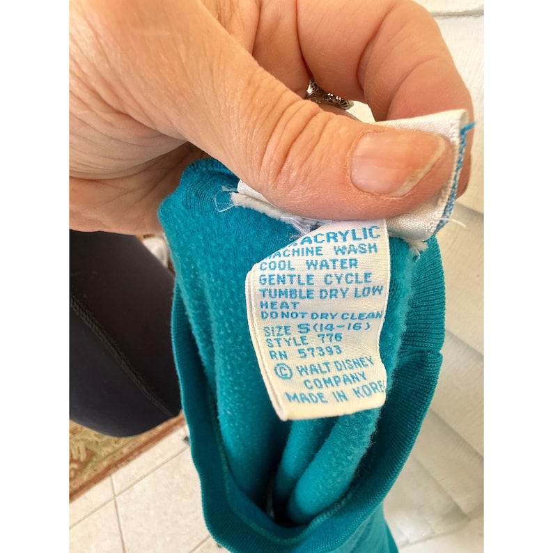 May include: A close-up of a teal acrylic garment tag with white text. The tag provides washing instructions, size, style, and RN information. The tag also includes the Walt Disney Company logo and states "MADE IN KORE".