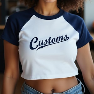 Woman at a major league baseball game wearing a custom baseball style cropped raglan baby tee with pinstripes in a design that would be great for the New York Yankees, Cleveland Guardians, Detroit Tigers, Houston Astros, Kansas City Royals, Los Angeles Angels, Seattle Mariners, St Louis Cardinals, Tampa Bay Rays, Toronto Blue Jays, Reno Aces, Round Rock Express, Somerset Patriots, Durham Bulls, St. Paul Saints, Round Rock Express, Syracuse Mets, Lehigh Valley IronPigs, and the San Jose Giants.