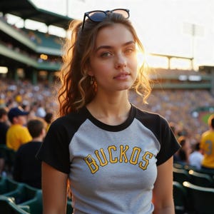 Woman at a MLB game rocking a custom baseball style cropped raglan baby tee with black sleeves and yellow writing that says Buccos.  The design could  work for the Pittsburgh Pirates, Akron RubberDucks, Salt Lake Bees, Charleston RiverDogs, Bowling Green Hot Rods, Rome Emperors, Myrtle Beach Pelicans, Brooklyn Cyclones, Altoona Curve, Bradenton Marauders, Rochester Red Wings, and San Jose Giants.