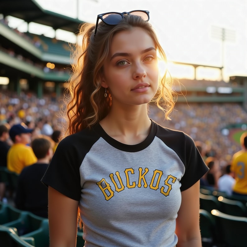 Woman at a MLB game rocking a custom baseball style cropped raglan baby tee with black sleeves and yellow writing that says Buccos.  The design could  work for the Pittsburgh Pirates, Akron RubberDucks, Salt Lake Bees, Charleston RiverDogs, Bowling Green Hot Rods, Rome Emperors, Myrtle Beach Pelicans, Brooklyn Cyclones, Altoona Curve, Bradenton Marauders, Rochester Red Wings, and San Jose Giants.