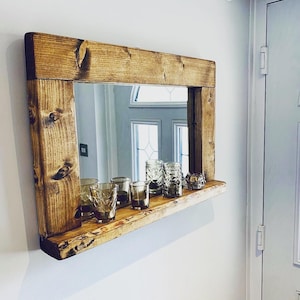 May include: A wooden framed mirror with a natural finish, mounted on a white wall. The mirror has a shelf holding several clear glass candle holders and drinking glasses. The mirror's frame is made of thick, square-cut timber.
