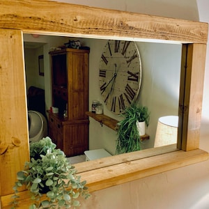 May include: A rectangular mirror with a wooden frame. The frame is made of natural-finish wood. The mirror reflects a room with a large clock, wooden furniture, and a potted plant. The frame is made of thick, unfinished wood.