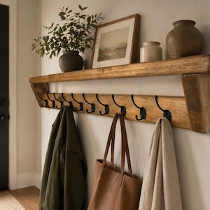 May include: A wooden wall shelf with black hooks below. A framed landscape picture, a small jar, and a brown vase are on the shelf. A green jacket, a brown tote bag, and a beige towel hang from the hooks.