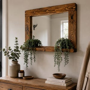 May include: A rectangular wall mirror featuring a dark stained wooden frame with a prominent grain pattern and an integrated narrow shelf at the bottom. The mirror reflects a room interior and is adorned with two trailing green plants.