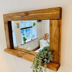 May include: A rectangular wooden mirror with a natural finish, mounted on a white wall. The mirror's frame is made of thick, stained wood. A small potted plant hangs below the mirror. The mirror reflects a room with a window.