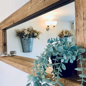 May include: A wooden framed mirror with a shelf. A black potted plant with trailing green leaves sits on the shelf. Two glass candle holders and a metal bucket with dried flowers are also visible. The mirror reflects a light fixture.
