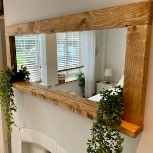 May include: A rectangular wall mirror featuring a thick, natural-toned wooden frame with visible grain and knots. The mirror includes an integrated wooden shelf along the bottom edge, which holds two trailing green artificial plants on each side.