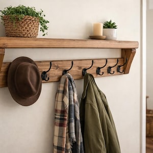 May include: A wooden wall-mounted shelf unit with six black metal hooks. A brown hat, a plaid scarf, and an olive green jacket hang from the hooks. A woven basket with greenery and a small potted plant sit on the shelf.