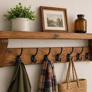 May include: A wooden wall shelf with six black metal hooks below. The shelf displays a framed picture, a brown glass bottle, and a potted plant. A green jacket, a plaid scarf, and a woven bag hang from the hooks.