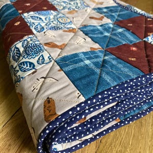 Puede incluir: Una colcha de patchwork con un patrón repetitivo de cuadrados en tonos rojo oscuro, azul y gris claro. Los cuadrados de tela muestran varios estampados, incluyendo motivos florales, cuadros y pequeñas botas de vaquero marrones. Borde azul oscuro.