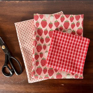 May include: Three folded pieces of fabric and black metal scissors. One fabric features a small red floral pattern on a light pink background, another displays a red strawberry print on light pink, and the third has a red and white gingham check.