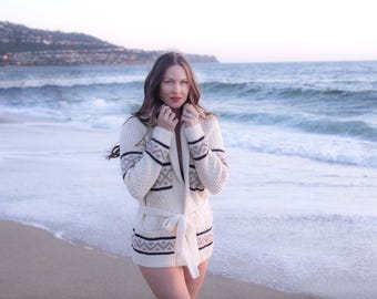 May include: A woman wearing a white cardigan with a black and brown geometric pattern on a beach. The cardigan has a tie belt and a collar.