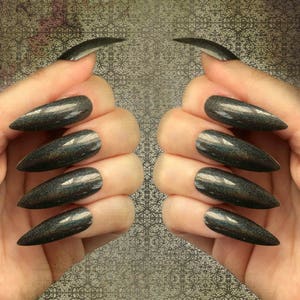 White female left and right hand wearing long black holographic stiletto press on nails on a textured gray background.