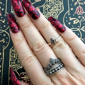 A white female hand is wearing extra long red coffin nails with black geometric detailing on an old book with a red, black and gold cover. the female hand is wearing several silver and black rings.