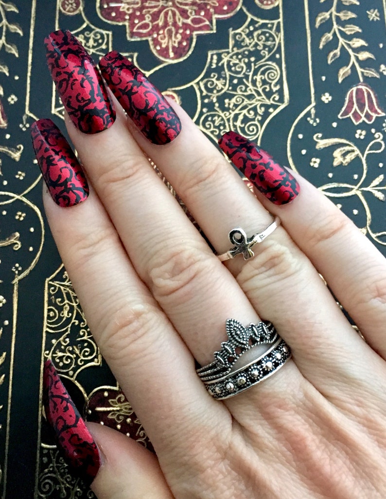 A white female hand is wearing extra long red coffin nails with black geometric detailing on an old book with a red, black and gold cover. the female hand is wearing several silver and black rings.