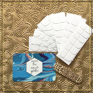 A stack of double sided nail tabs is fanned out over a gold background, next to a small file and branded sticker. Also called jelly gummy glue or nail tape, this item is used to apply artificial nails.