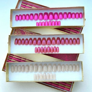 Three rectangular boxes contain individual sets of pink press on nails in square, oval and coffin shapes on  white background.