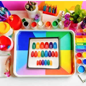 A square box of 20 rainbow colored almond press on nails is plced on a colorful paint tray on a desk with paints and chalk surrounding it.