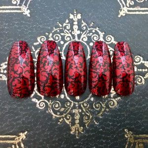 Close up of 5 long red coffin false nails with black geometric detailing on black background with gold geometric detail.
