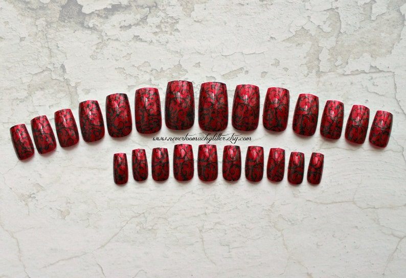 a full set of 20 metallic red press on nails in square shape with black geometric detailing on a faded white background.