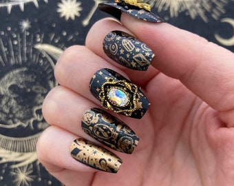 May include: Close-up of a hand wearing black and gold press-on nails with a celestial design. Each nail features a different design with stars, moons, and other celestial elements. One nail has a gold accent with a clear rhinestone.
