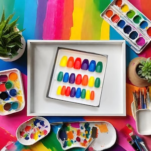 A square box of 20 rainbow colored coffin press on nails is placed on a colorful paint tray on a desk with paints and plants surrounding it.