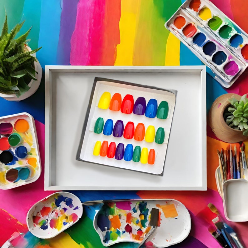 A square box of 20 rainbow colored coffin press on nails is placed on a colorful paint tray on a desk with paints and plants surrounding it.