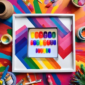 A square box of 20 rainbow colored square press on nails is placed on a colorful paint tray on a desk with paints and art supplies surrounding it.