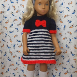 Könnte beinhalten: Eine Puppe trägt ein blau-weiß gestreiftes Kleid mit einer roten Schleife. Das Kleid hat einen roten Saum. Die Puppe trägt weiße Socken und schwarze Schuhe. Die Puppe steht vor einem weißen Hintergrund mit einem Muster aus blauen und orangefarbenen Segelbooten.