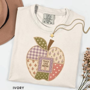 May include: An ivory t-shirt featuring a central apple-shaped applique made of various patterned fabric patches in shades of purple, green, and pink. The center patch displays the text "TEACH LOVE INSPIRE" with a small heart icon below.
