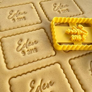 Peut inclure: Gros plan sur un emporte-pièce jaune avec le texte "Eden 9 ans" dessus. L'emporte-pièce est posé sur une feuille de pâte à biscuits avec plusieurs biscuits déjà découpés. Les biscuits sont également estampillés du texte "Eden 9 ans".