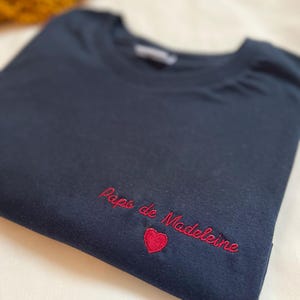 May include: A dark blue t-shirt with red embroidered text that says "Papa de Madeleine" with a red heart.