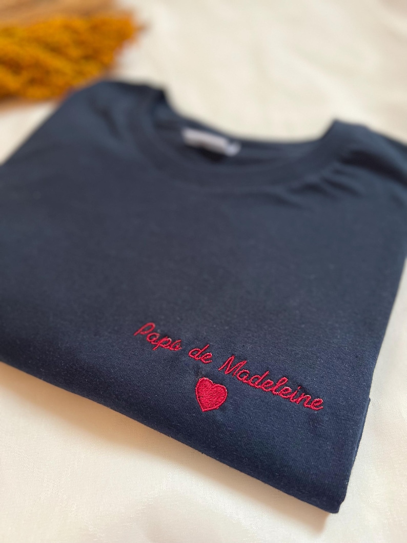 May include: A dark blue t-shirt with red embroidered text that says "Papa de Madeleine" with a red heart.