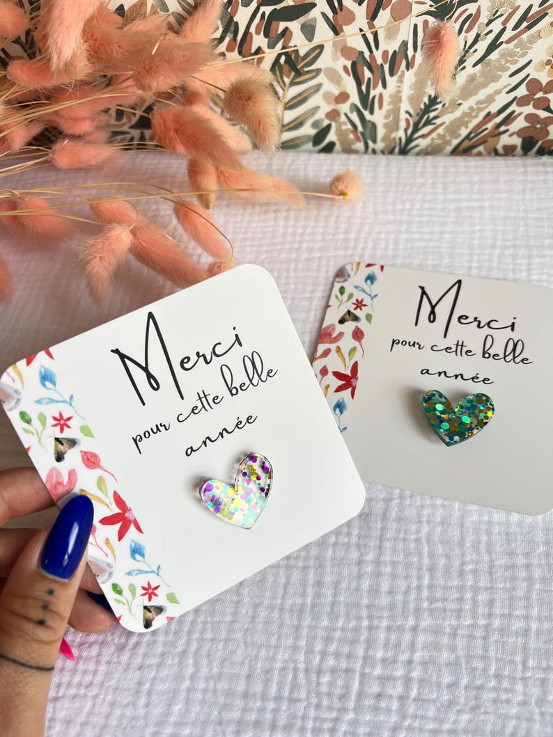 May include: Two white cards with floral designs and the text "Merci pour cette belle ann&eacute;e" (Thank you for this beautiful year) with a glittery heart on each card. The heart on the left card is iridescent and the heart on the right card is green.