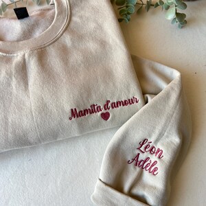 Op de afbeelding: Een beige sweatshirt met de tekst "Mamita d'amour" geborduurd op de voorkant en een hart. De mouw heeft de namen "Leon Adele" geborduurd.