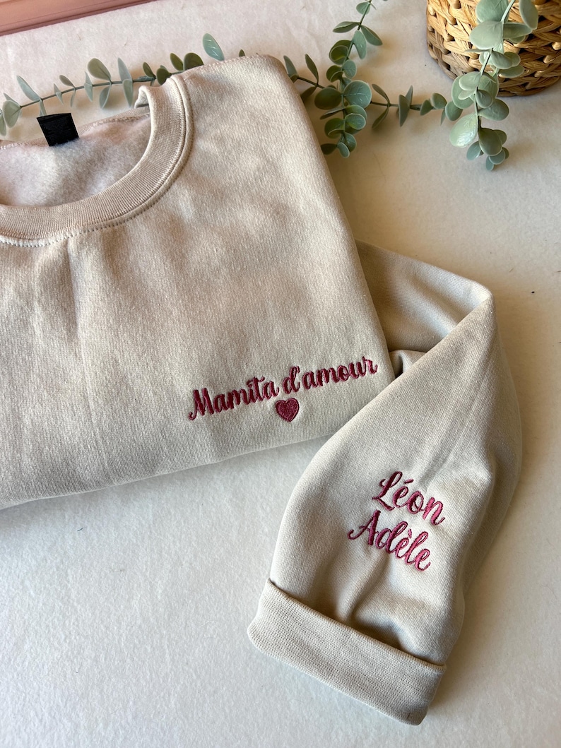 Op de afbeelding: Een beige sweatshirt met de tekst "Mamita d'amour" geborduurd op de voorkant en een hart. De mouw heeft de namen "Leon Adele" geborduurd.