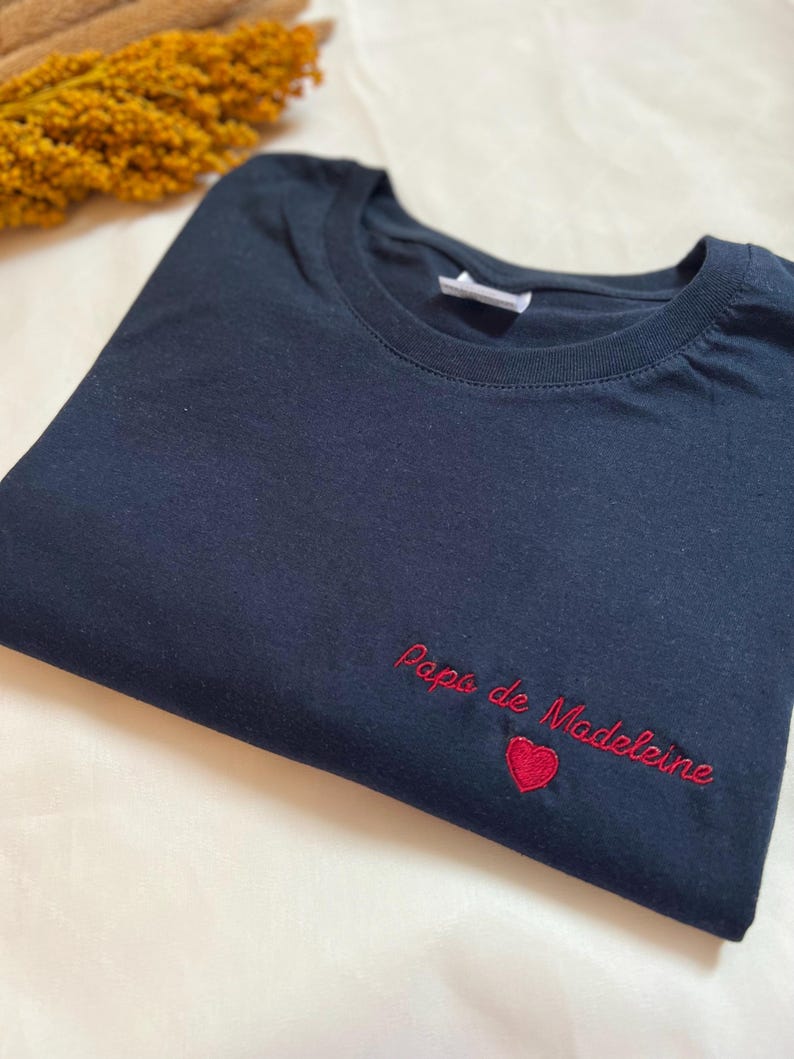 May include: A dark blue t-shirt with red embroidery that says "Papa de Madeleine" with a small red heart.