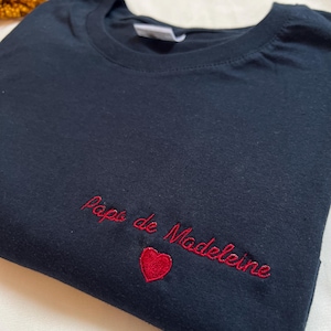 May include: A black t-shirt with red embroidery that says "Papa de Madeleine" with a red heart.