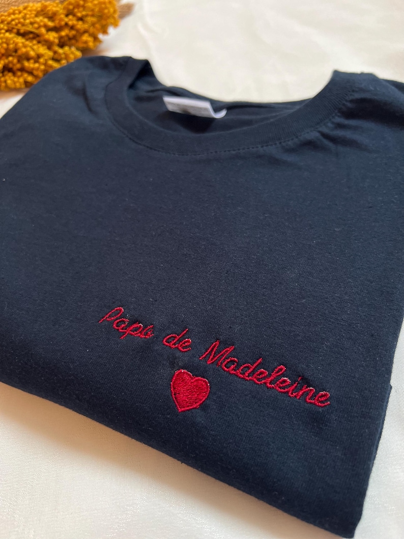 May include: A black t-shirt with red embroidery that says "Papa de Madeleine" with a red heart.