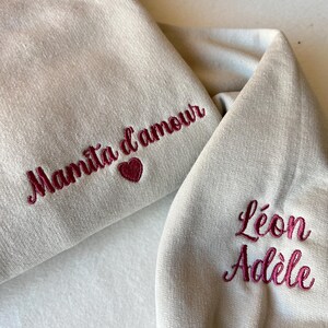 Op de afbeelding: Twee beige sweatshirts met geborduurde tekst. E&eacute;n sweatshirt zegt "Mamita d'amour" met een hart. De andere sweatshirt zegt "L&eacute;on Ad&egrave;le".