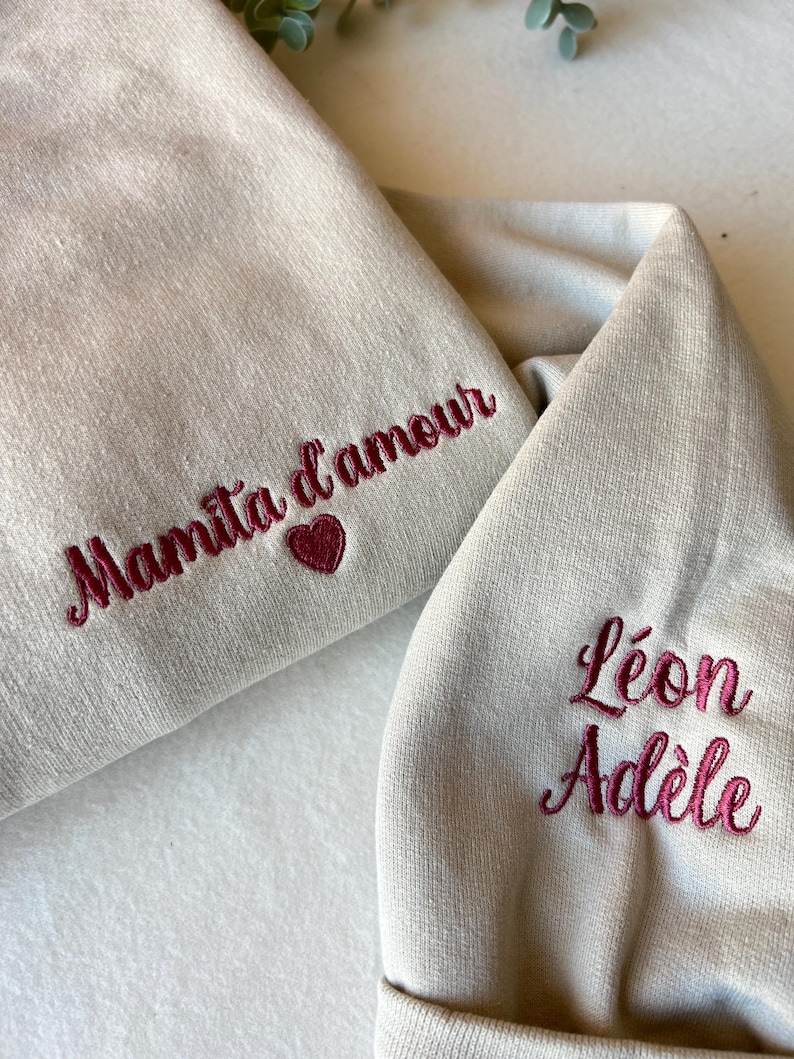 Op de afbeelding: Twee beige sweatshirts met geborduurde tekst. E&eacute;n sweatshirt zegt "Mamita d'amour" met een hart. De andere sweatshirt zegt "L&eacute;on Ad&egrave;le".