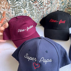 Peut inclure: Trois casquettes de baseball avec du texte brodé. Une casquette est bordeaux avec du texte blanc qui dit "père-fection". Une autre casquette est noire avec du texte rouge qui dit "père-fect". La troisième casquette est bleu marine avec du texte blanc qui dit "Super Papa" et un cœur rouge.