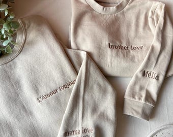 May include: Beige sweatshirts with embroidered text. One sweatshirt says "brother love" in brown thread. Another says "L'Amour toujours" and "mama love Maël & Mélio" in brown thread. The sleeves of the sweatshirts are folded.