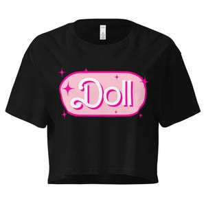 May include: Black crop top with a pink oval graphic that says "Doll" in a white script font. The graphic is outlined in pink and has pink starbursts on each side. The shirt has short sleeves.