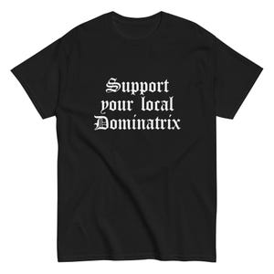 May include: Black t-shirt with the white text "Support your local Dominatrix" in a gothic font. The shirt is a crew neck style and appears to be made of a soft material. The text is centered on the front of the shirt.