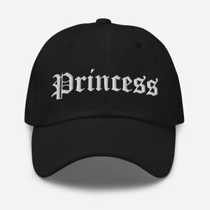 May include: Black baseball cap with white embroidered text that reads "Princess" in an old English font.