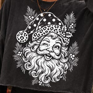 K&ouml;nnte beinhalten: Ein anthrazitfarbenes, lang&auml;rmeliges, bauchfreies Sweatshirt mit einem wei&szlig;en Grafikdruck eines zwinkernden Weihnachtsmanns, der eine M&uuml;tze mit Leopardenmuster tr&auml;gt, umgeben von Tannennadeln und Stechpalmenbeeren, mit einem rohen Saum am Bund.