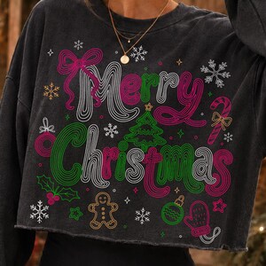 May include: A charcoal gray cropped sweatshirt featuring a graphic print with the text Merry Christmas in pink and green wavy lettering. The design includes festive icons like a gingerbread man, a candy cane, a mitten, and snowflakes.