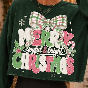 K&ouml;nnte beinhalten: Ein dunkelgr&uuml;nes, lang&auml;rmeliges bauchfreies Sweatshirt mit einem gro&szlig;en Grafikdruck einer karierten Schleife, einer Glocke und dem Text "MERRY joyful & bright CHRISTMAS" in Rot, Gr&uuml;n und Wei&szlig;. Das Design zeigt Stechpalmenbl&auml;tter und Schneeflocken.